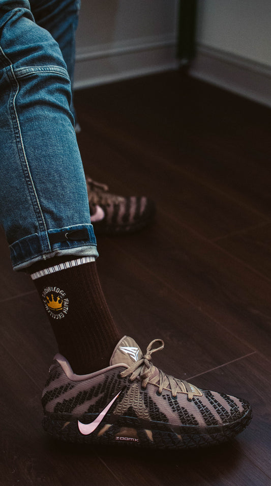 Person wearing 17th and Granville Black Excellence sock with a logo and a sneaker on a wooden floor.