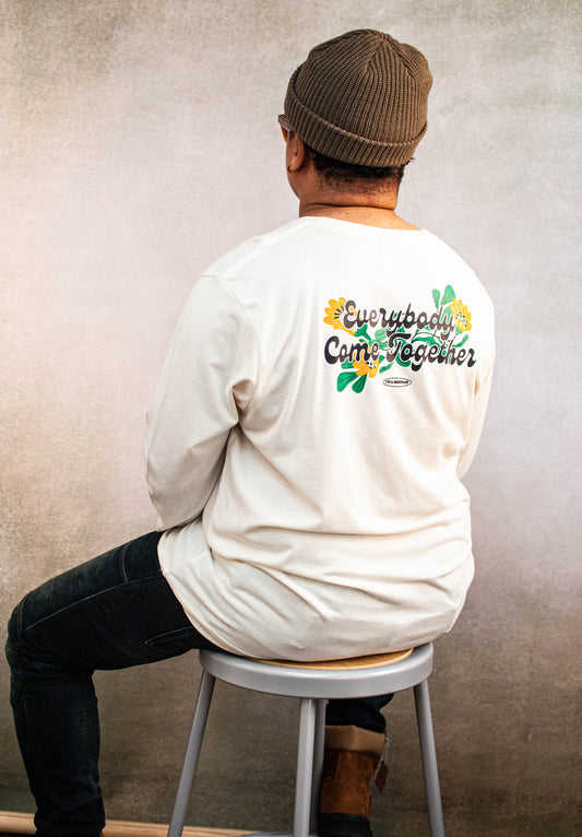 Person wearing a cream long-sleeve shirt with a colorful graphic highlight "Everybody Come Together" on the back, sitting on a stool against a plain wall.