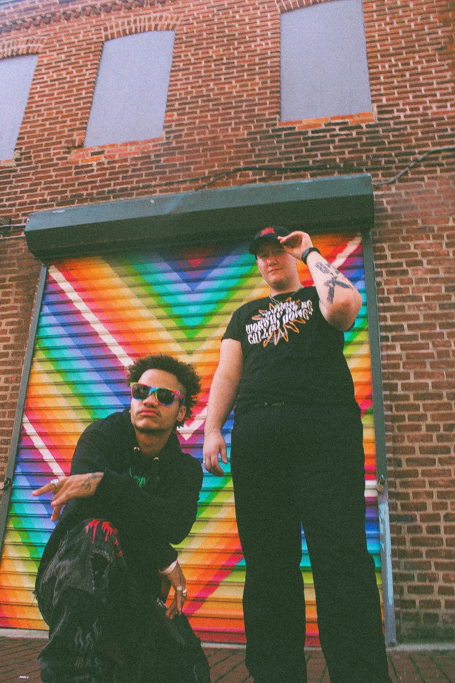 Two BIPOC LGBTQ+ models wearing Eintou Collection black hat and t-shirt, standing in front of colorful mural. 17th & Granville x C. Alexandria Bernard Thomas collaboration.