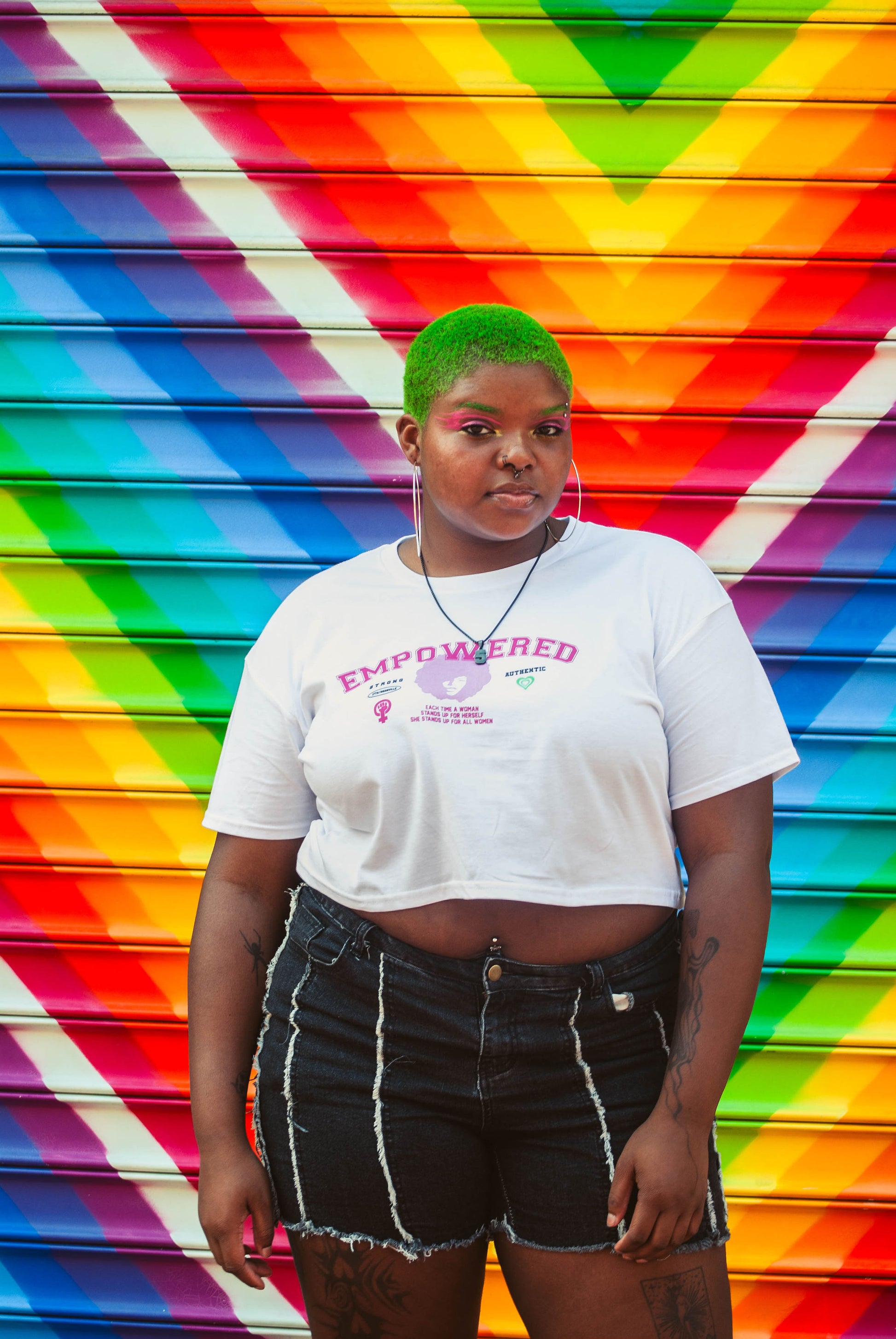 A person with neon green hair and a white Empowered Crop Top from 17th & Granville, a black-owned company, stands in front of a vibrant rainbow-striped wall. They are wearing denim shorts and large hoop earrings, exuding confidence.