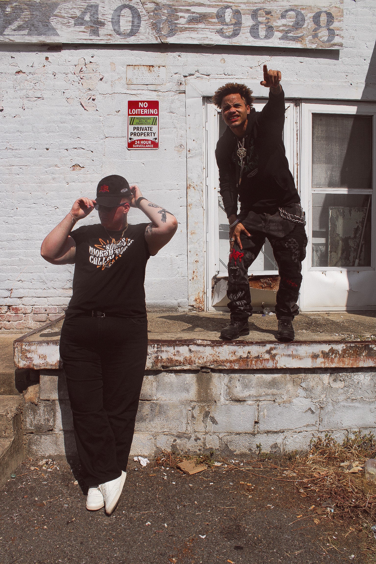 Two individuals stand on concrete steps in front of a brick wall. One leans casually, adjusting their cap and 17th & Granville poetry hoodie, while the other stands on the edge with one arm pointing upward. A No Loitering sign and a partial phone number are visible in the background.