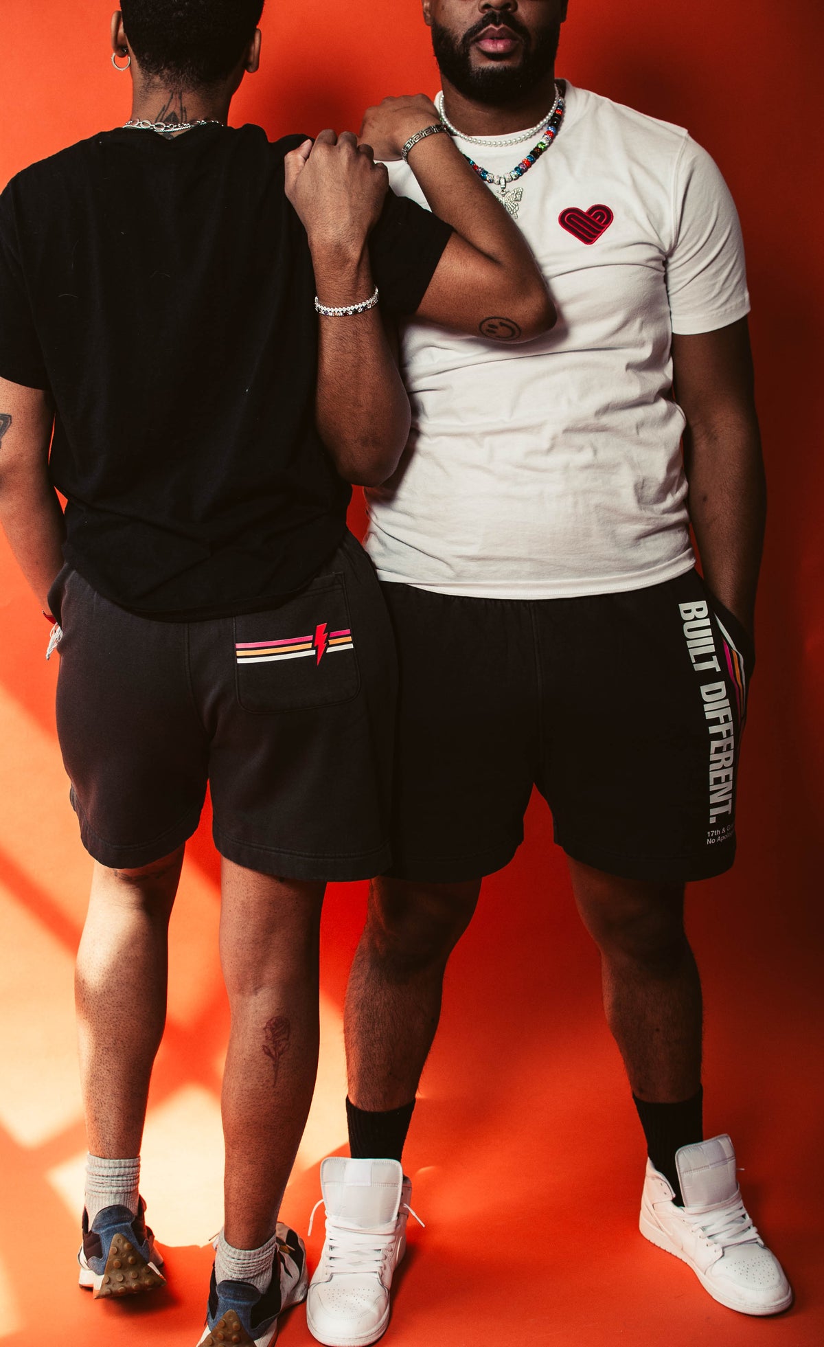 Two people standing against an orange background, wearing No Apologies shorts with visible branding.