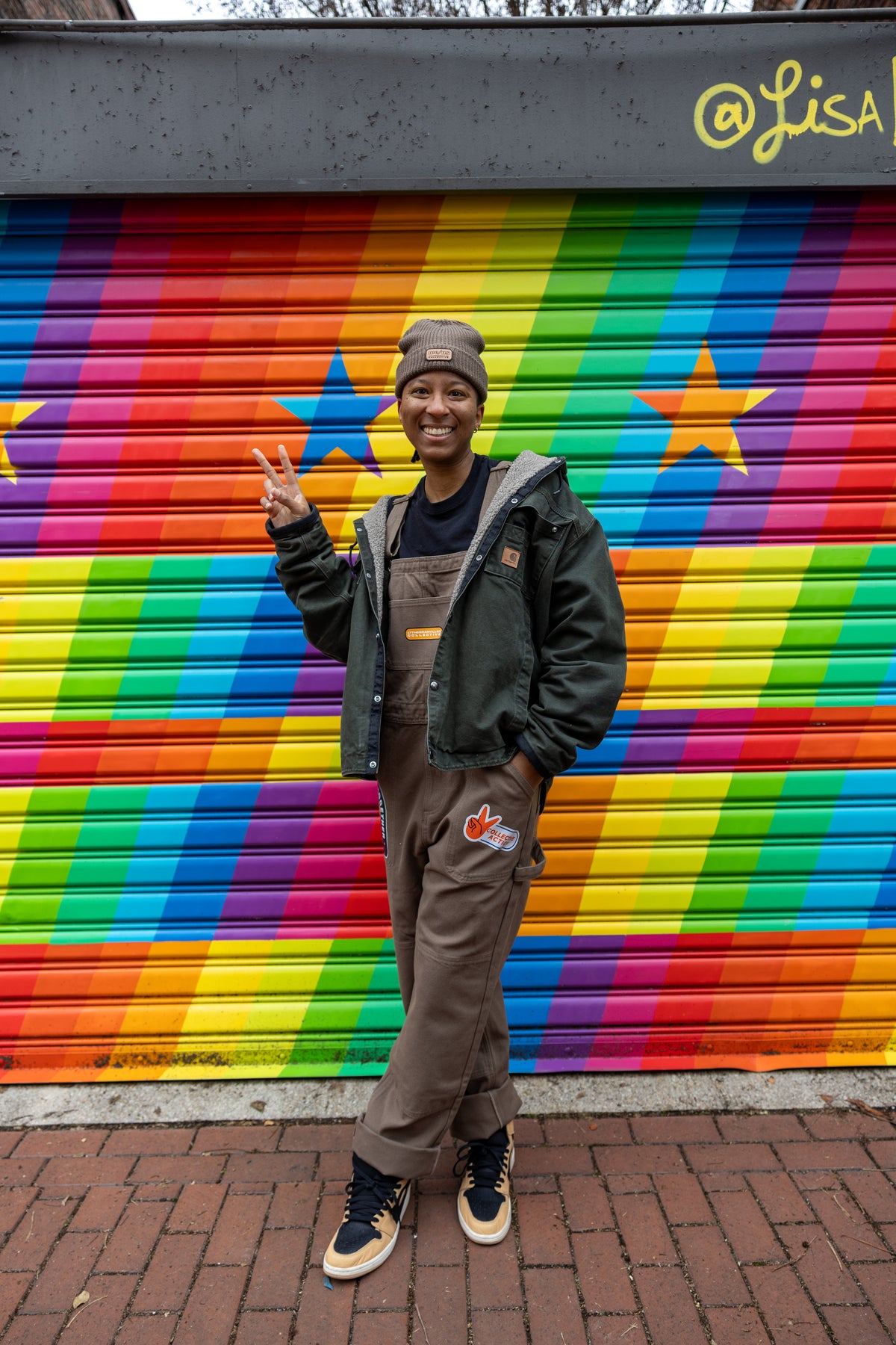 Model wearing brown We Us overalls with a We Us beanie in front of a rainbow background.