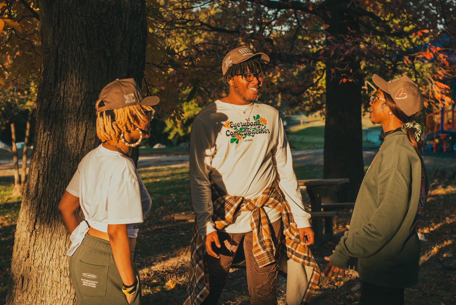 Three people in casual clothing and caps are standing and conversing in a sunlit park surrounded by trees. The person in the center wears a long-sleeved shirt with text, while the others are in short sleeves and a hoodie.