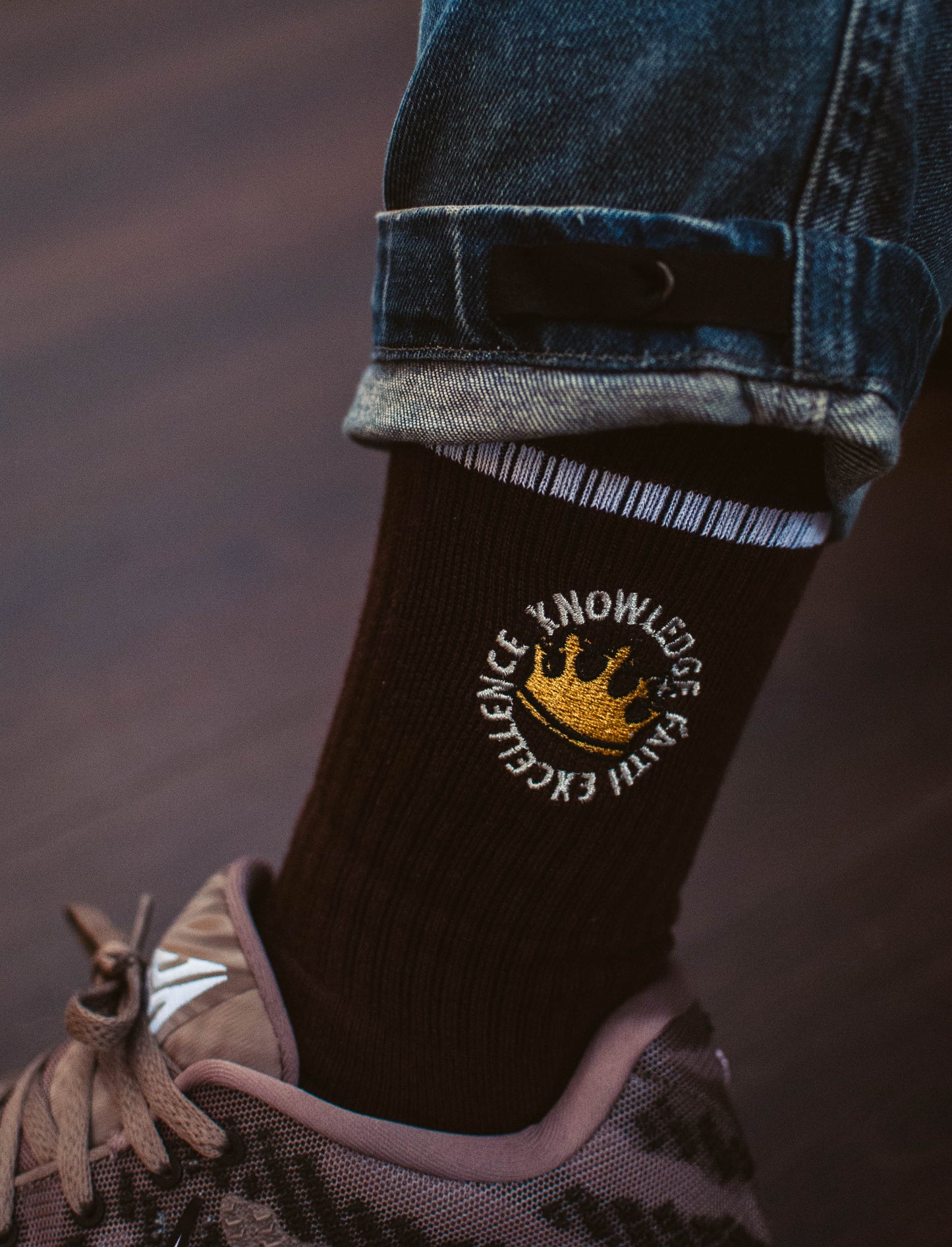 Espresso Brown sock with a logo of a crown and the words Knowledge, Faith, Excellence, worn by a person with rolled-up jeans