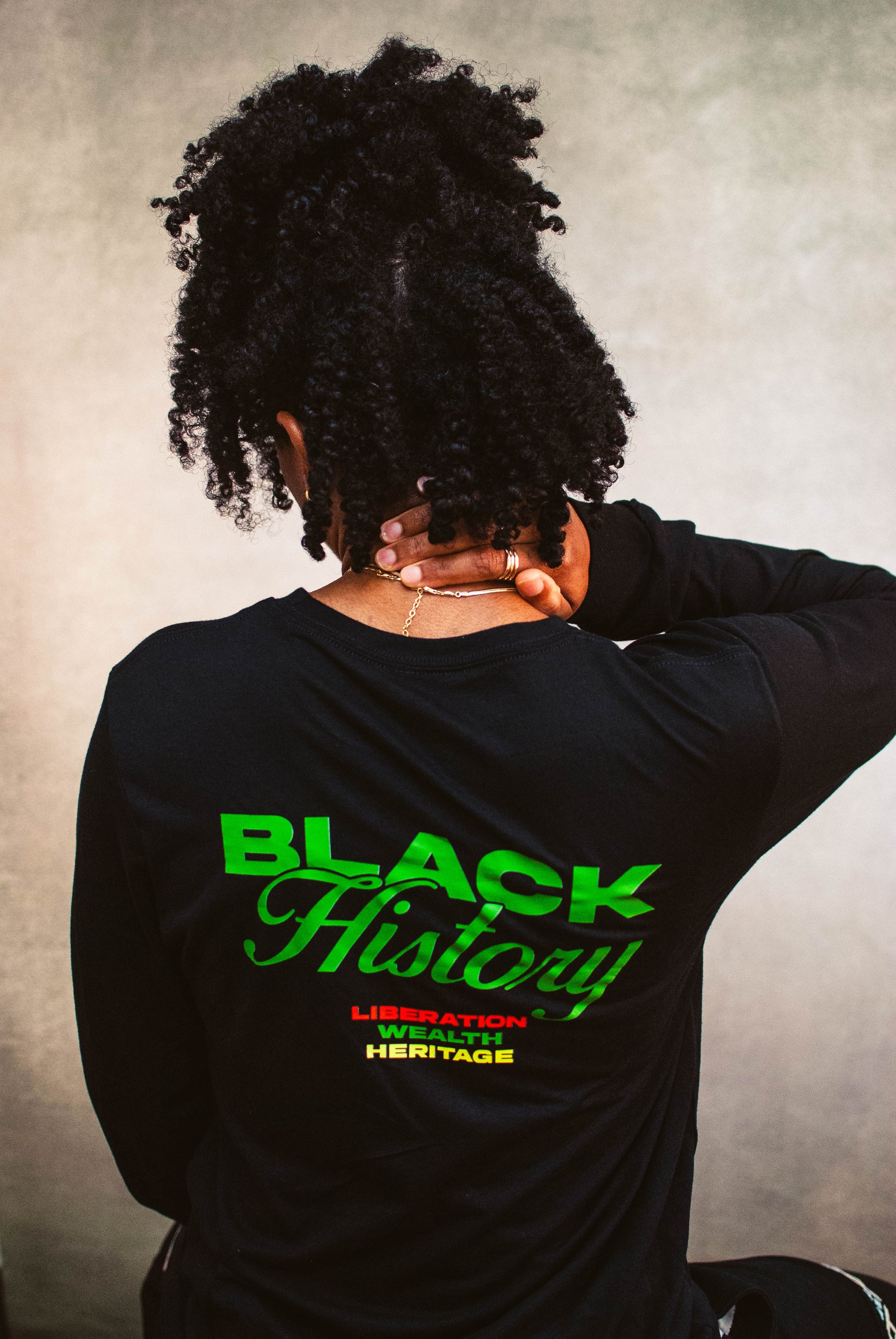 Person wearing a black long-sleeve shirt with 'Black History' text in green and yellow on the back, against a textured gray background.