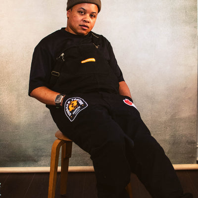 A person sitting on a chair with black overalls embellished with patches on each pocket and one on the chest., with a brown beanie.
