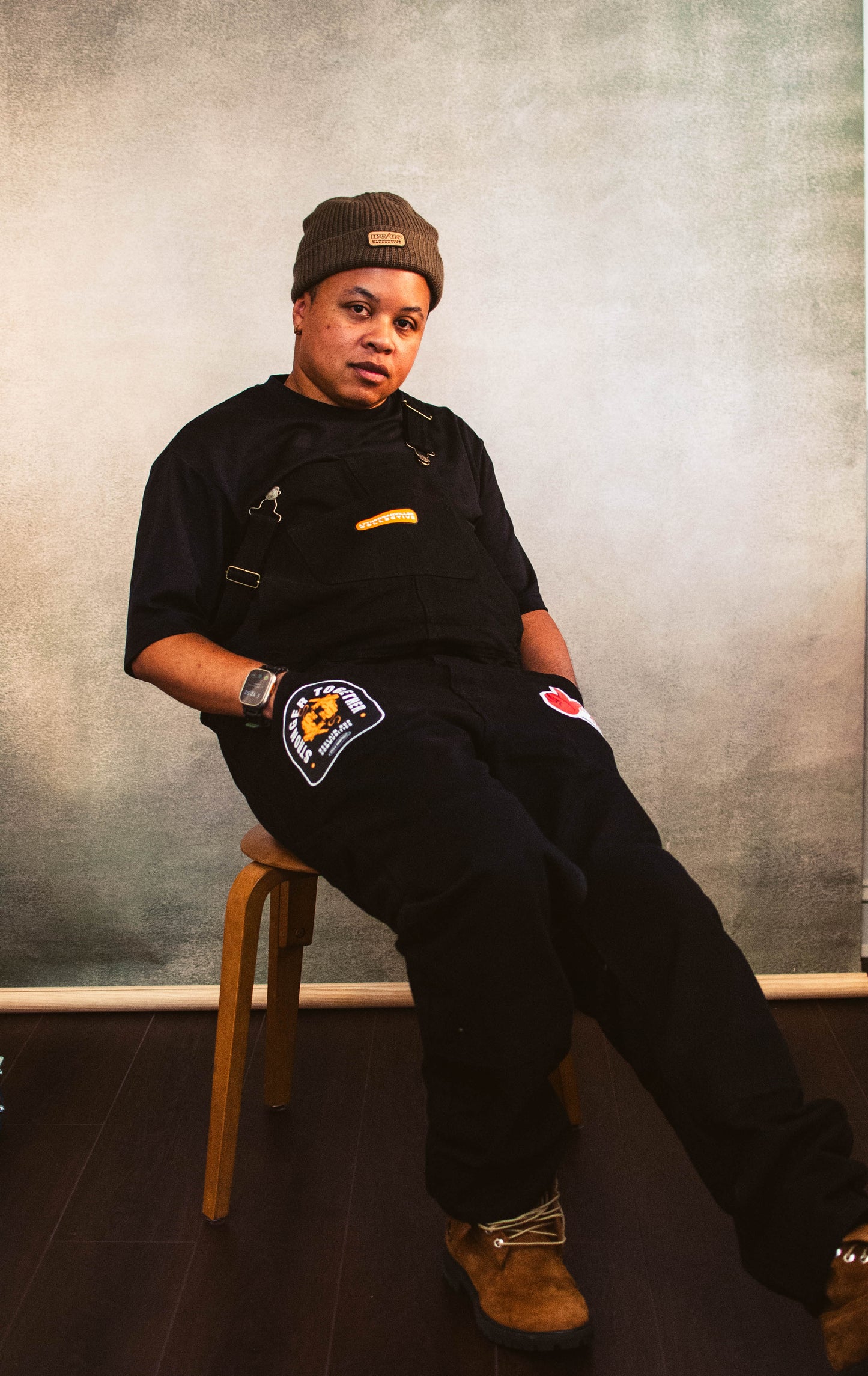 A person sitting on a chair with black overalls embellished with patches on each pocket and one on the chest., with a brown beanie.
