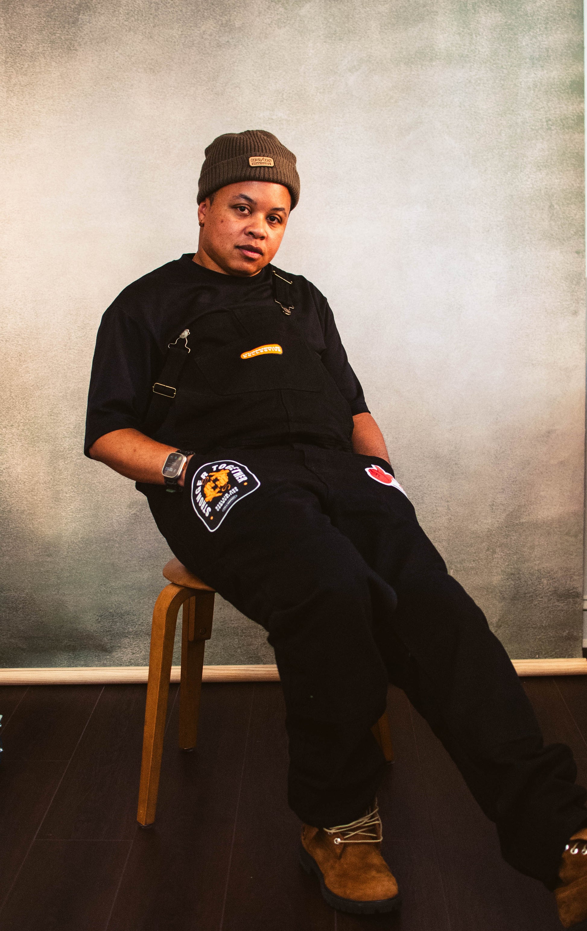 A person sitting on a chair with black overalls embellished with patches on each pocket and one on the chest., with a brown beanie.