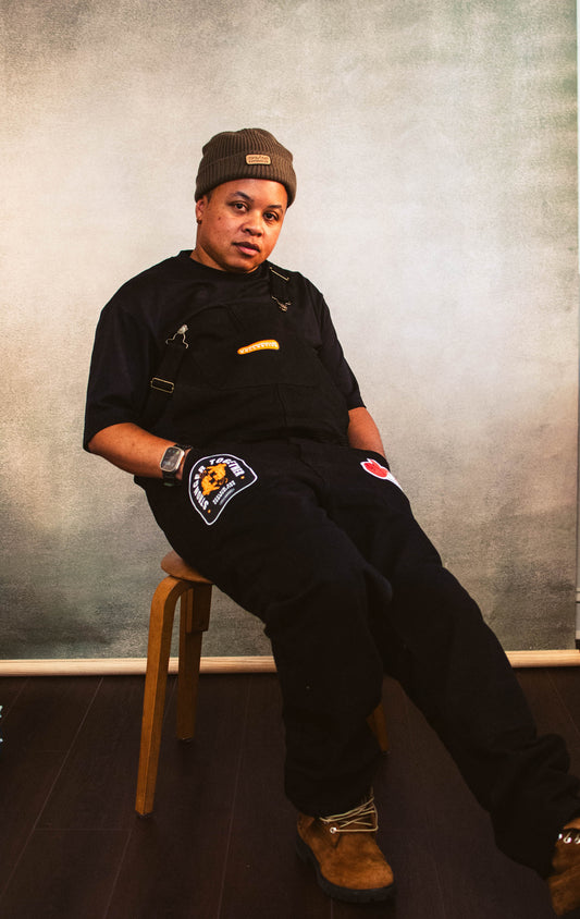 A person sitting on a chair with black overalls embellished with patches on each pocket and one on the chest., with a brown beanie.