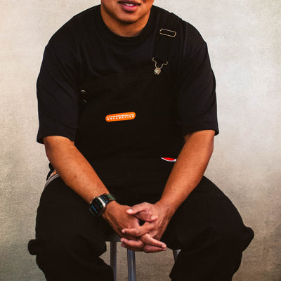 Person wearing a black shirt and brown beanie sitting on a stool against a plain background