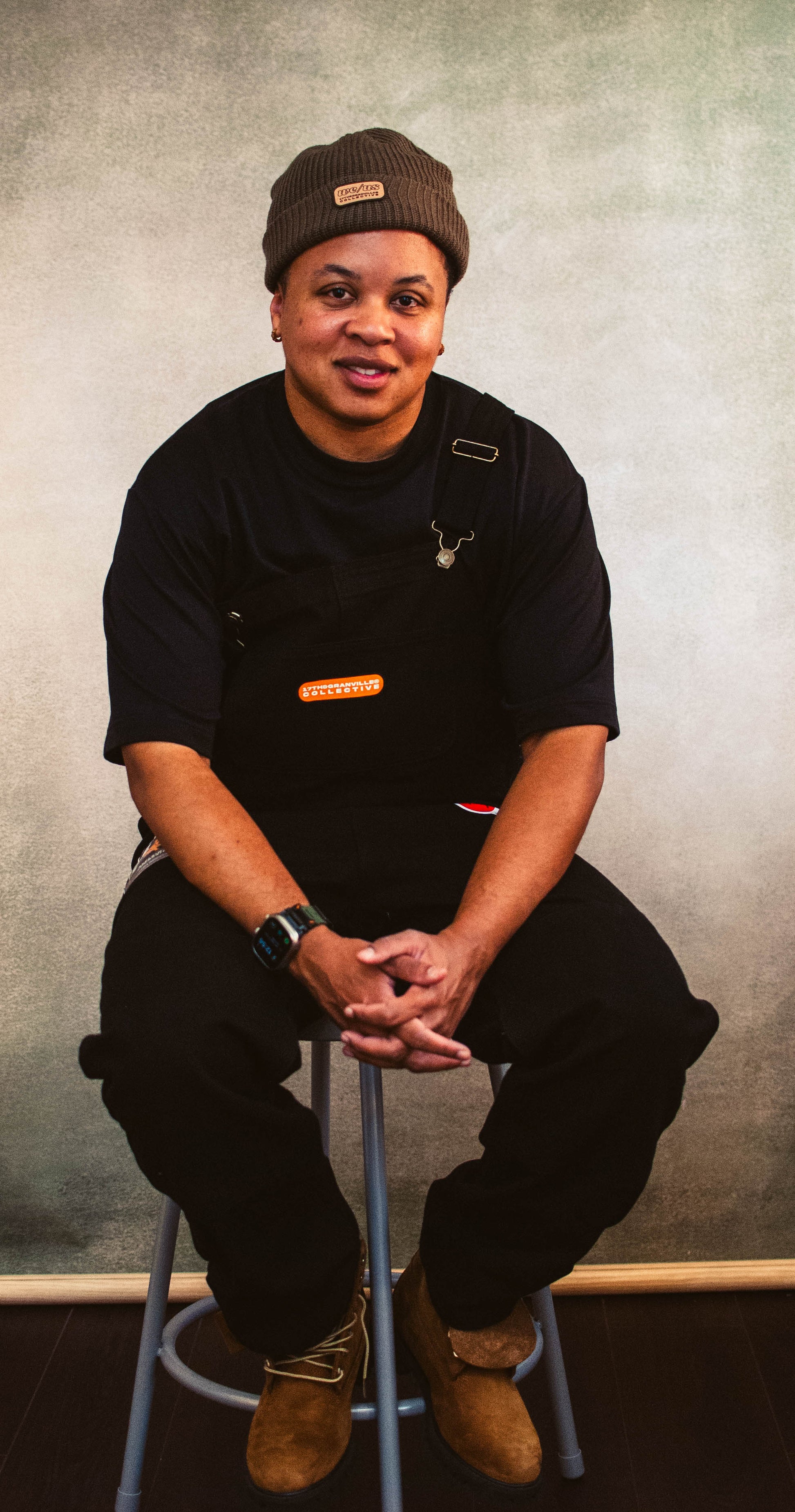 Person wearing a black shirt and brown beanie sitting on a stool against a plain background
