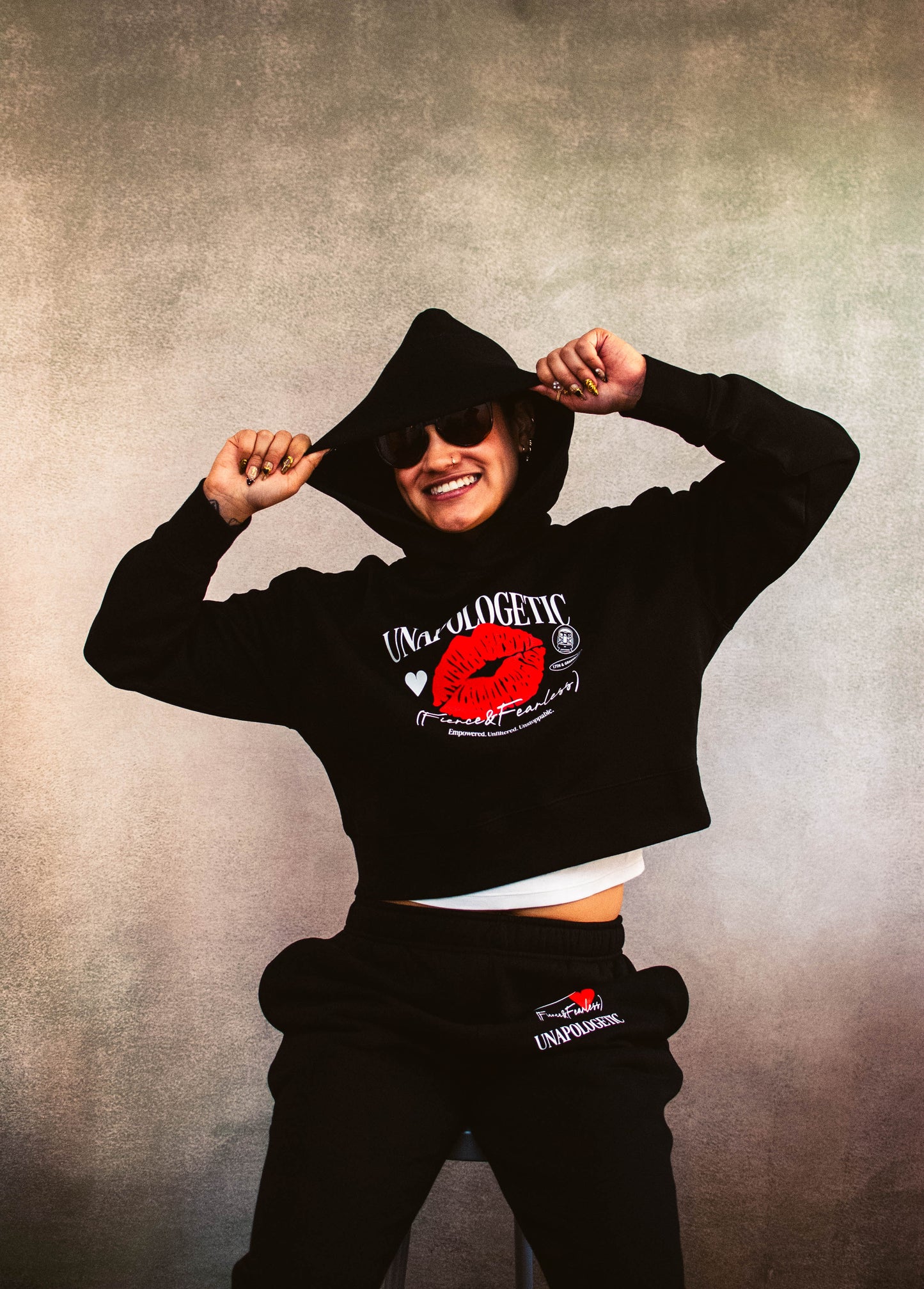 Person wearing a black crop hoodie and sweatpants with an unapologetic, fierce and fearless emblem design against a textured gray background