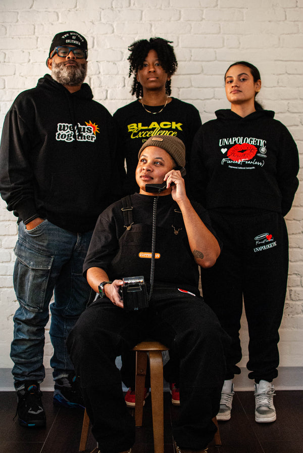Four individuals wearing black hoodies and apparel from 17th and Granville with various designs: WE/US, Feminine Energy, Black Excellence against a white brick wall.
