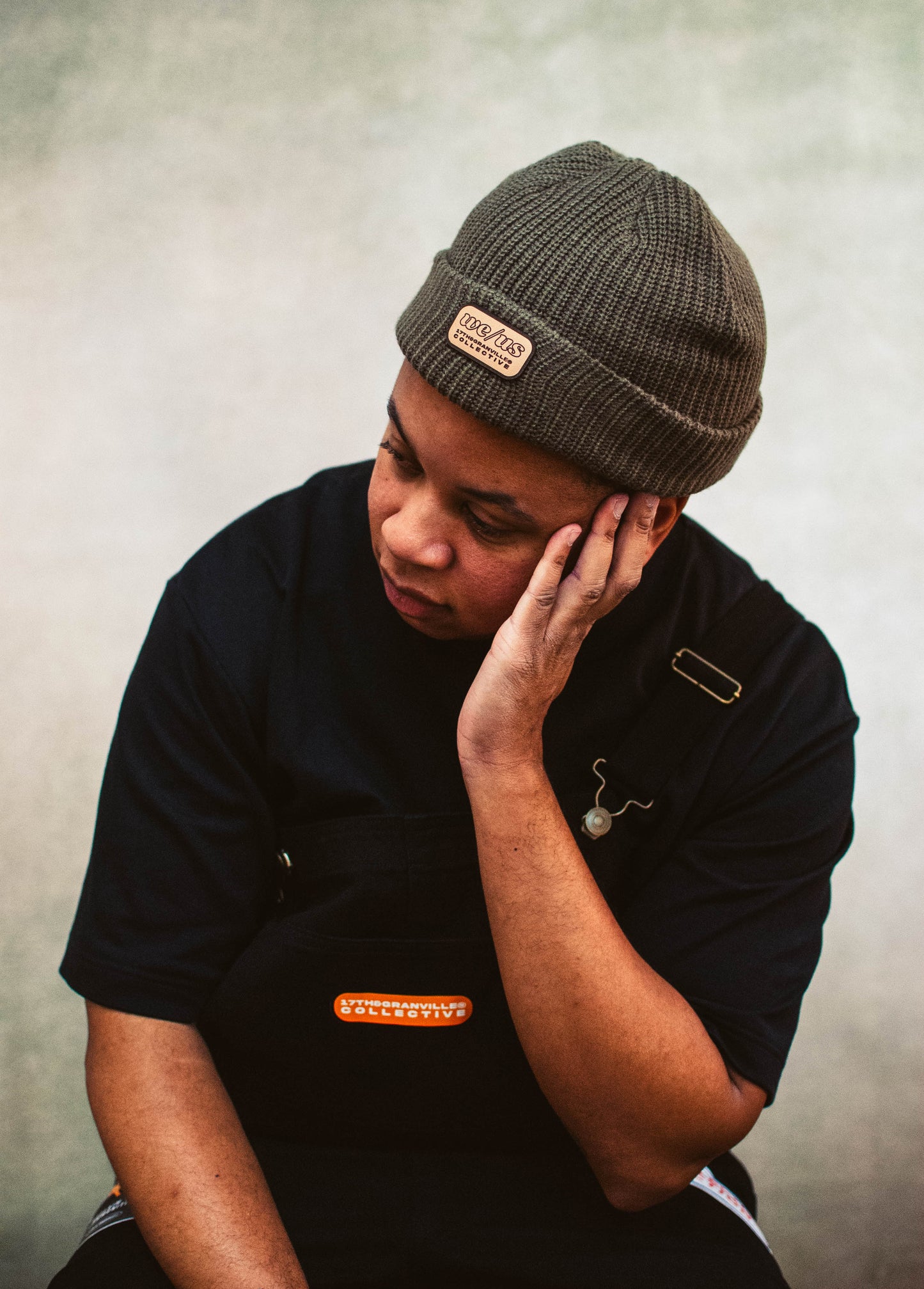 A person with their hand on their head with a brown beanie, black shirt and black overalls