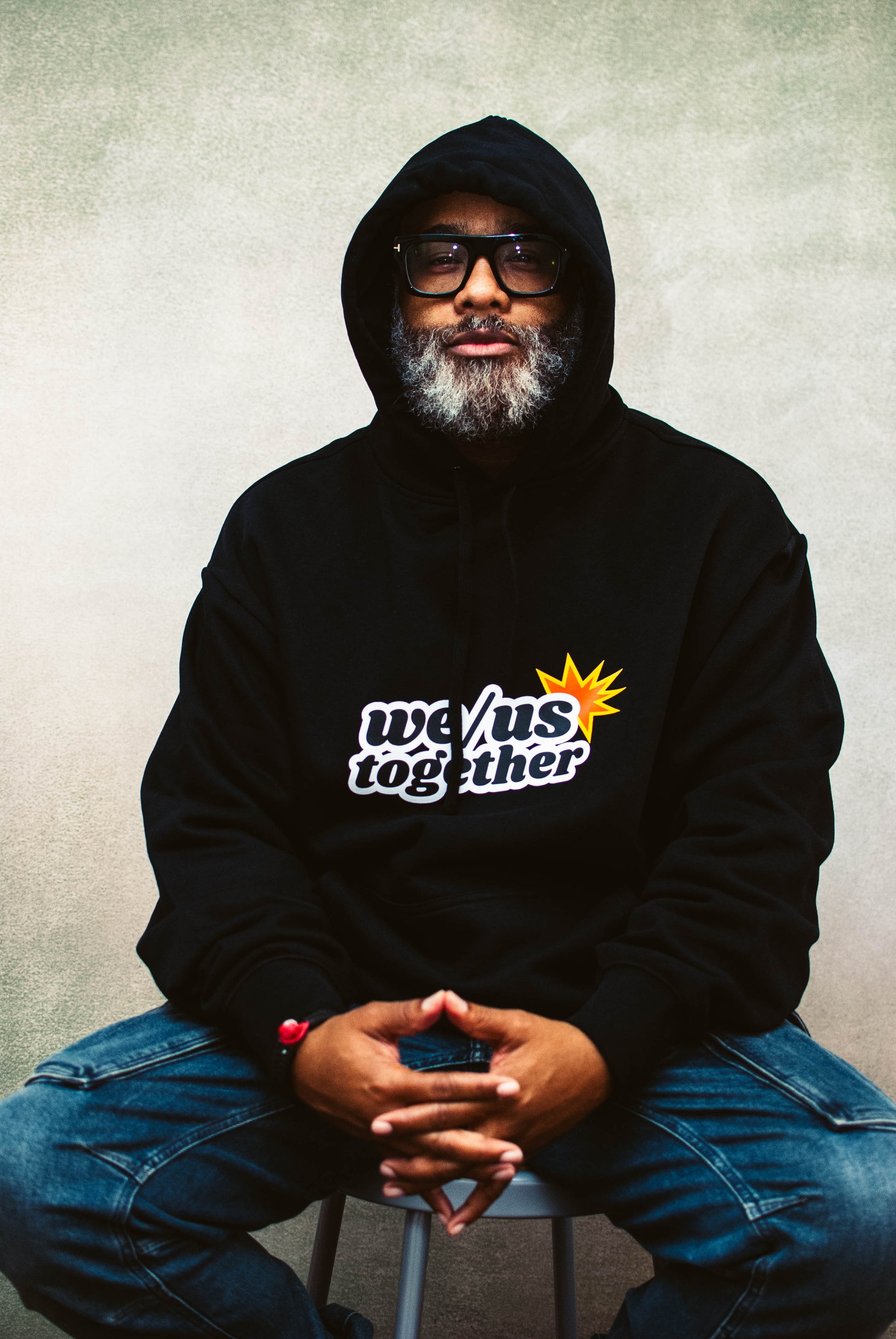 Person wearing a black hoodie with 'we/us together' text, sitting on a stool against a plain background