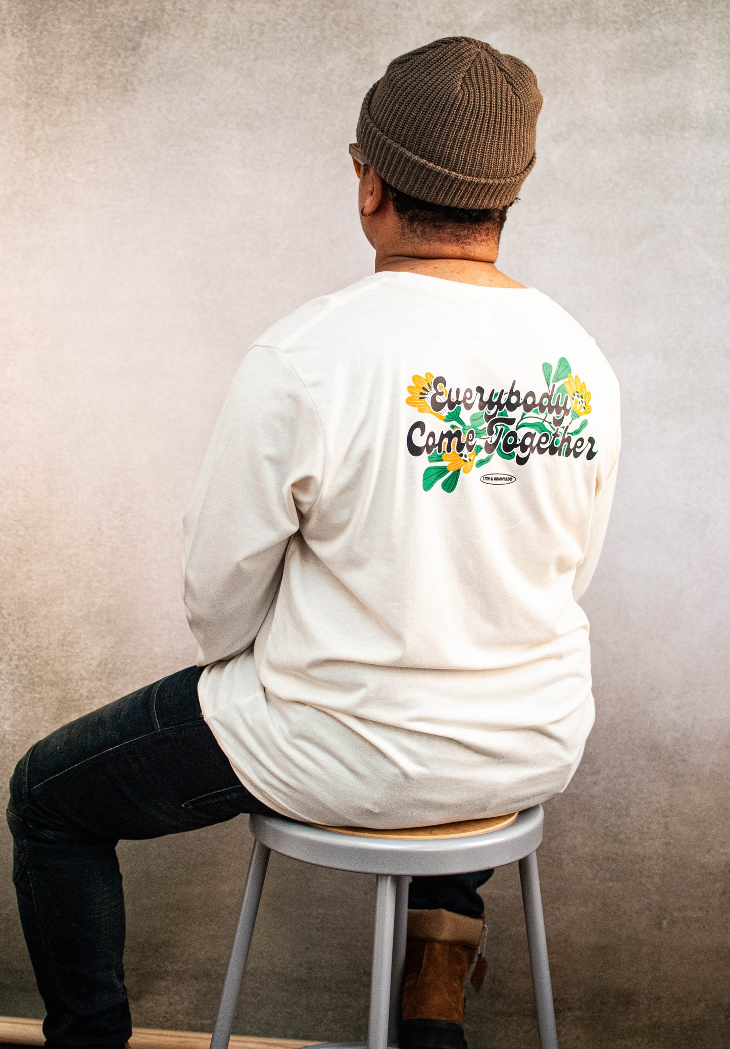 Person wearing a cream long-sleeve shirt with a colorful graphic highlight "Everybody Come Together" on the back, sitting on a stool against a plain wall.
