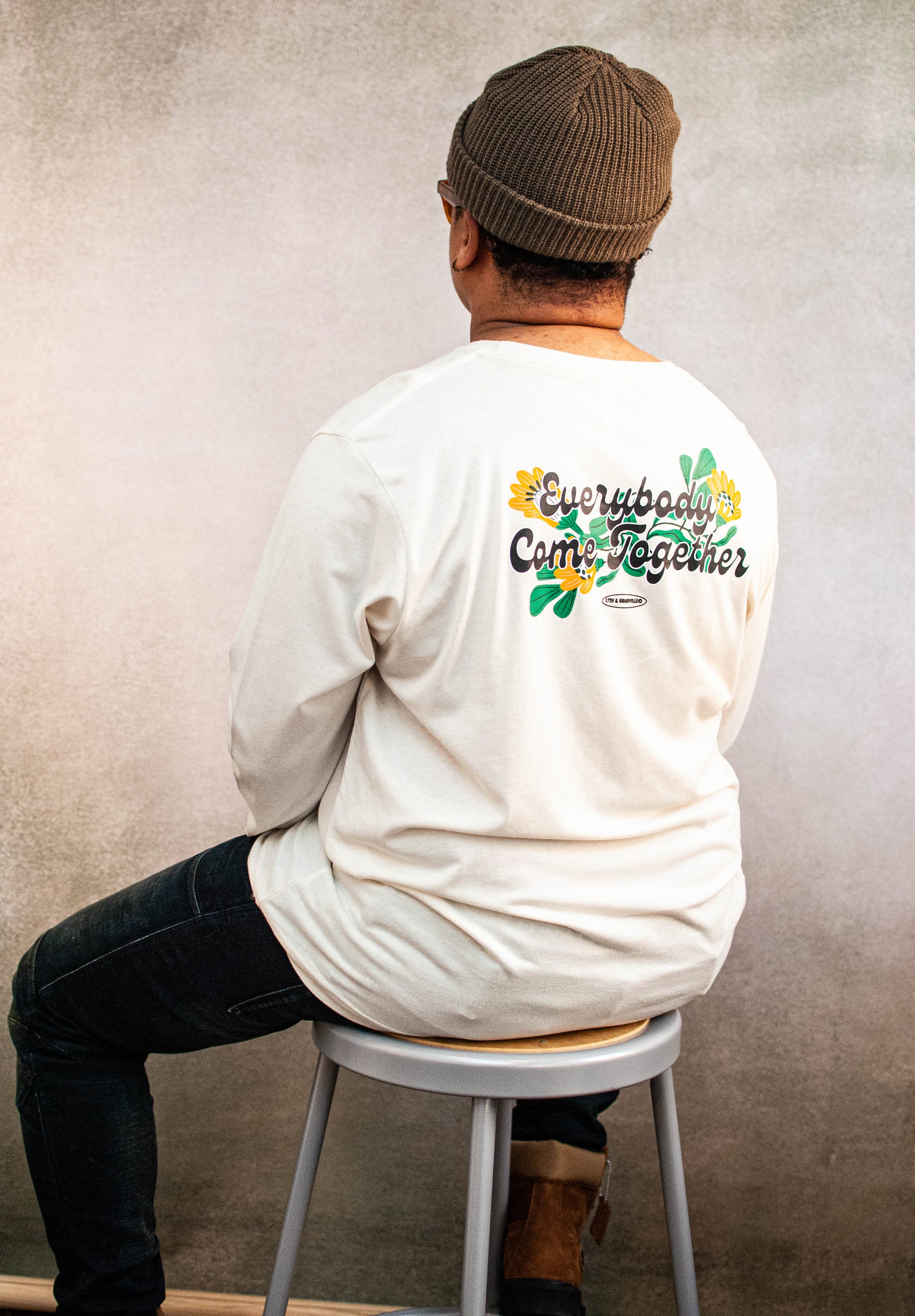 Person wearing a cream long-sleeve shirt with a colorful graphic highlight "Everybody Come Together" on the back, sitting on a stool against a plain wall.