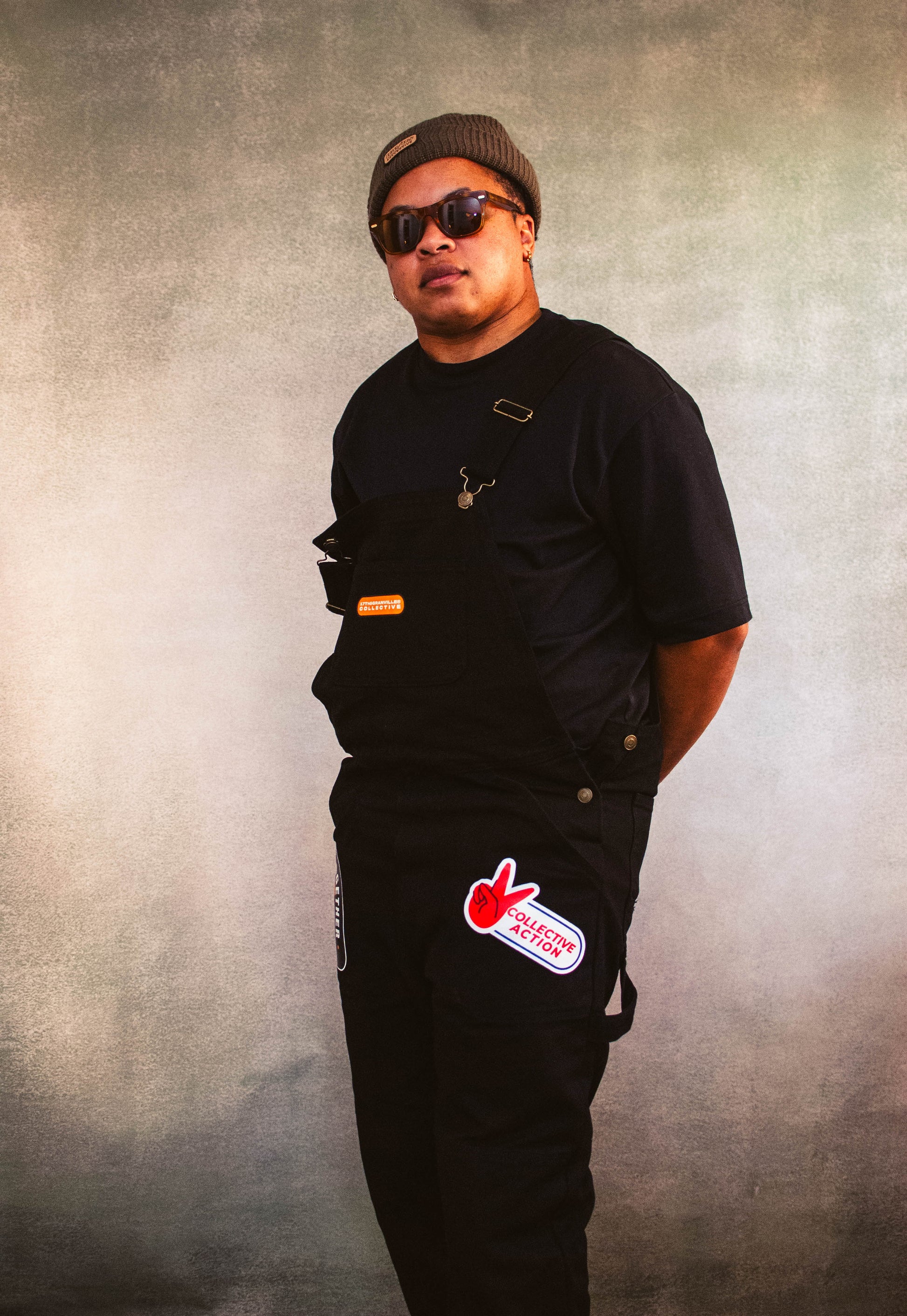 Person wearing 17th and Granville black overalls with a collective action logo on pants against a textured gray background
