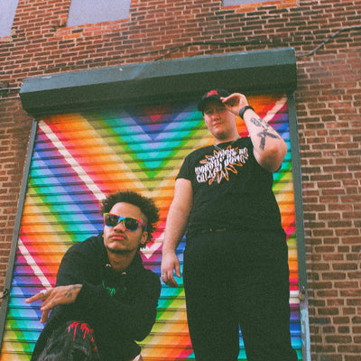 Two BIPOC LGBTQ+ models wearing Eintou Collection black hat and t-shirt, standing in front of colorful mural. 17th & Granville x C. Alexandria Bernard Thomas collaboration.