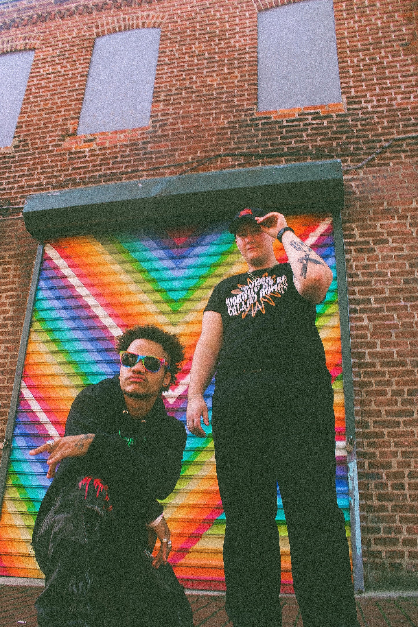 Two BIPOC LGBTQ+ models wearing Eintou Collection black hat and t-shirt, standing in front of colorful mural. 17th &amp; Granville x C. Alexandria Bernard Thomas collaboration.