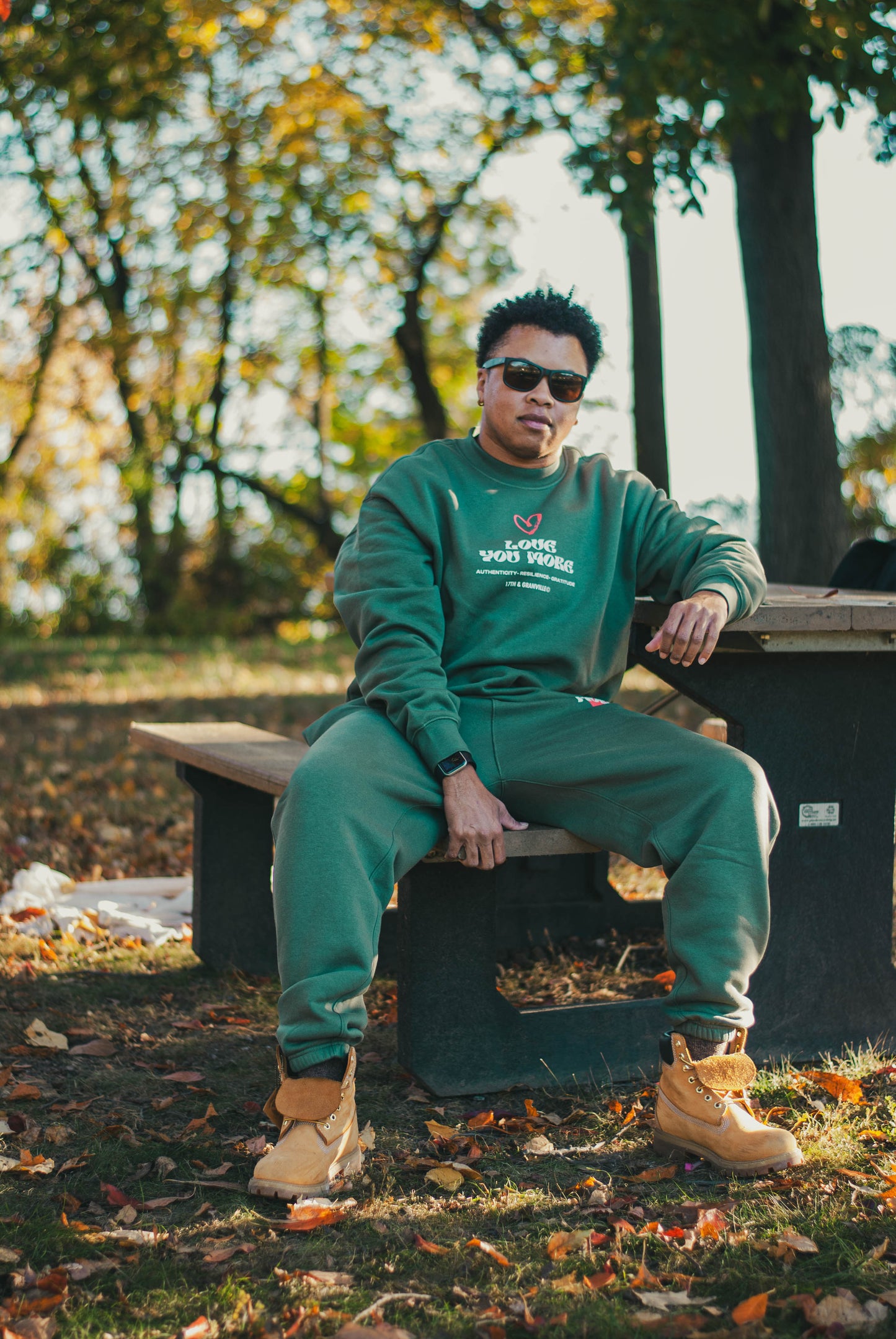 A person wearing a 17th & Granville green tracksuit, along with sunglasses and tan boots, relaxes on a picnic table in a park. Celebrating self-expression among autumn trees adorned with golden leaves on the ground, this scene embodies the perfect fall essential setting.