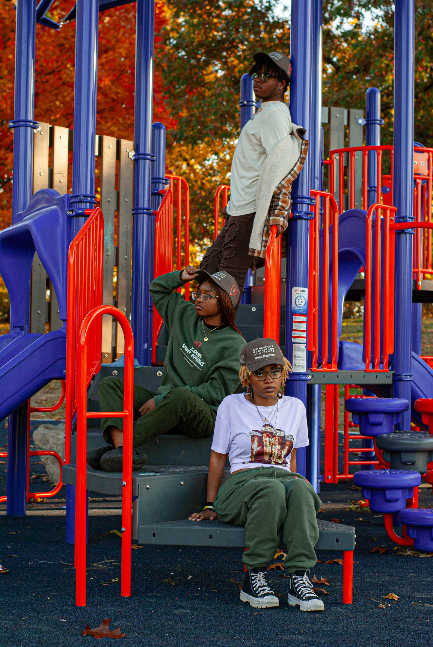 Three individuals express themselves on a lively playground structure featuring red and blue elements. They are dressed in casual attire, accessorized with hats, glasses, and an eye-catching "Love You More" sweatshirt by 17th & Granville, amidst the vivid autumn leaves in the background.