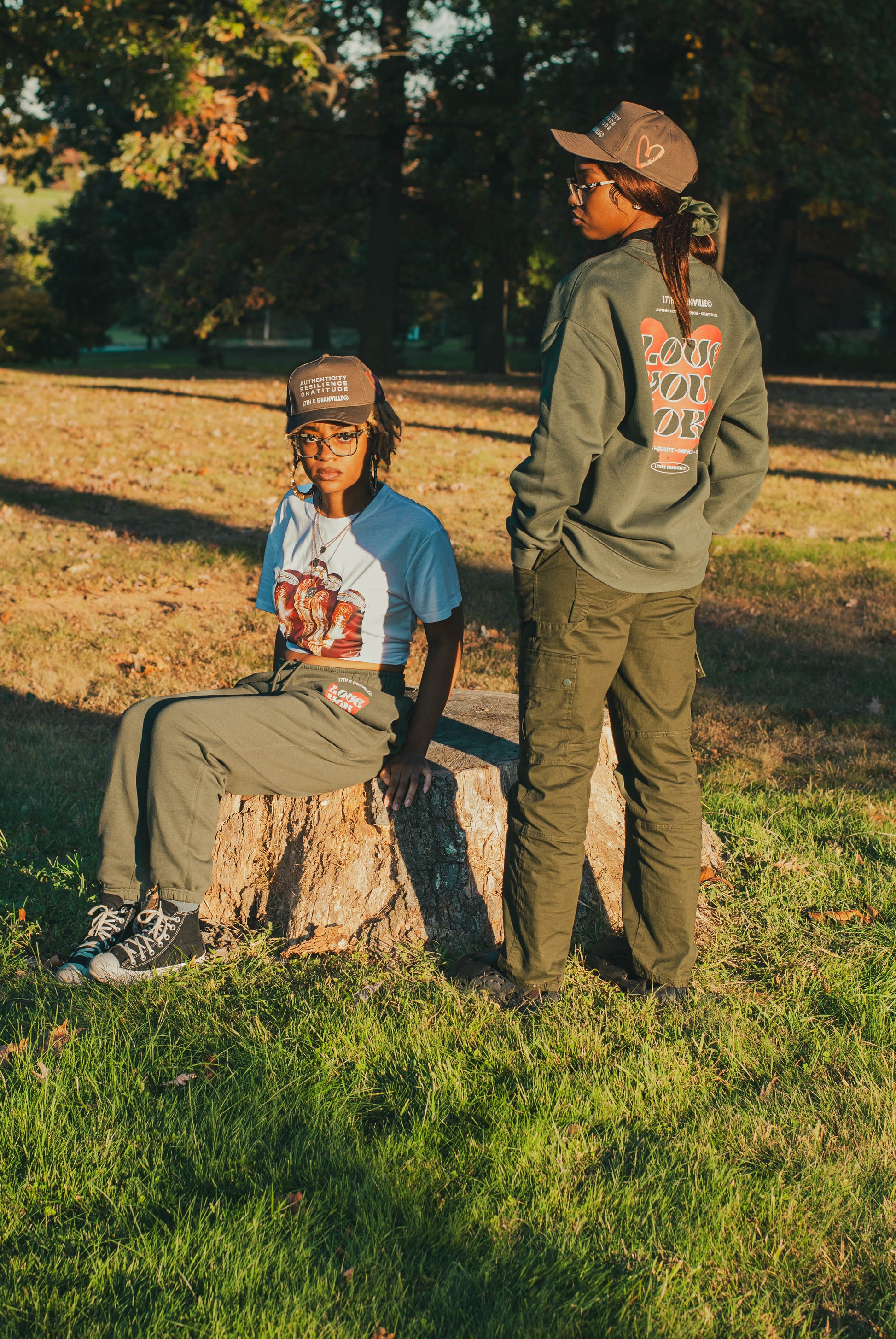 Two people are outdoors in a park. One sits confidently on a tree stump wearing glasses, a graphic tee, and green pants, embodying self-expression. The other stands out in sunglasses, a cap, and an olive hoodie with text on the back—a quintessential fall piece from 17th & Granville. Both enjoy the warmth of the sunny day on the grass.