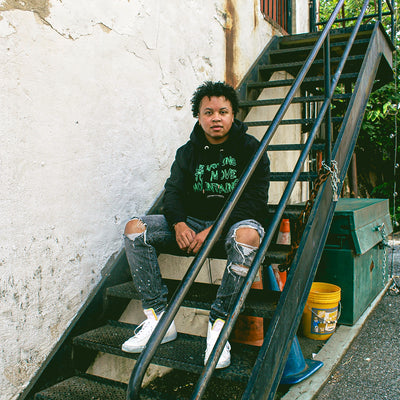 A Black model wearing the Eintou Collection's organic black hoodie with embroidered poetry excerpts, paired with a matching hat and t-shirt, showcasing 17th & Granville's collaboration with C. Alexandria Bernard Thomas in support of BIPOC and LGBTQ+ communities through fashion activism.