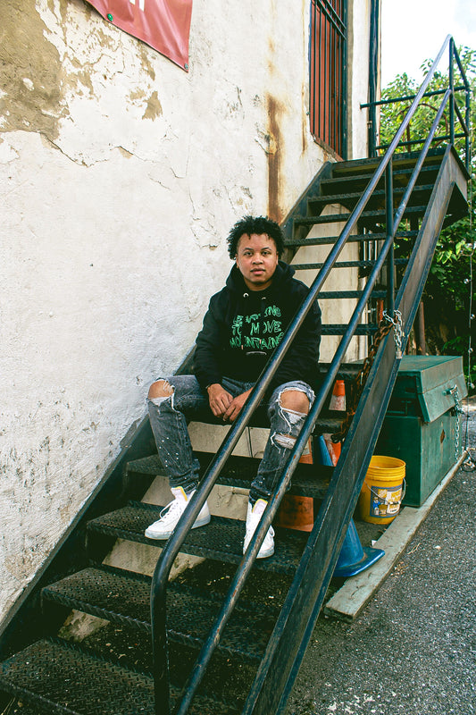 A Black model wearing the Eintou Collection's organic black hoodie with embroidered poetry excerpts, paired with a matching hat and t-shirt, showcasing 17th & Granville's collaboration with C. Alexandria Bernard Thomas in support of BIPOC and LGBTQ+ communities through fashion activism.