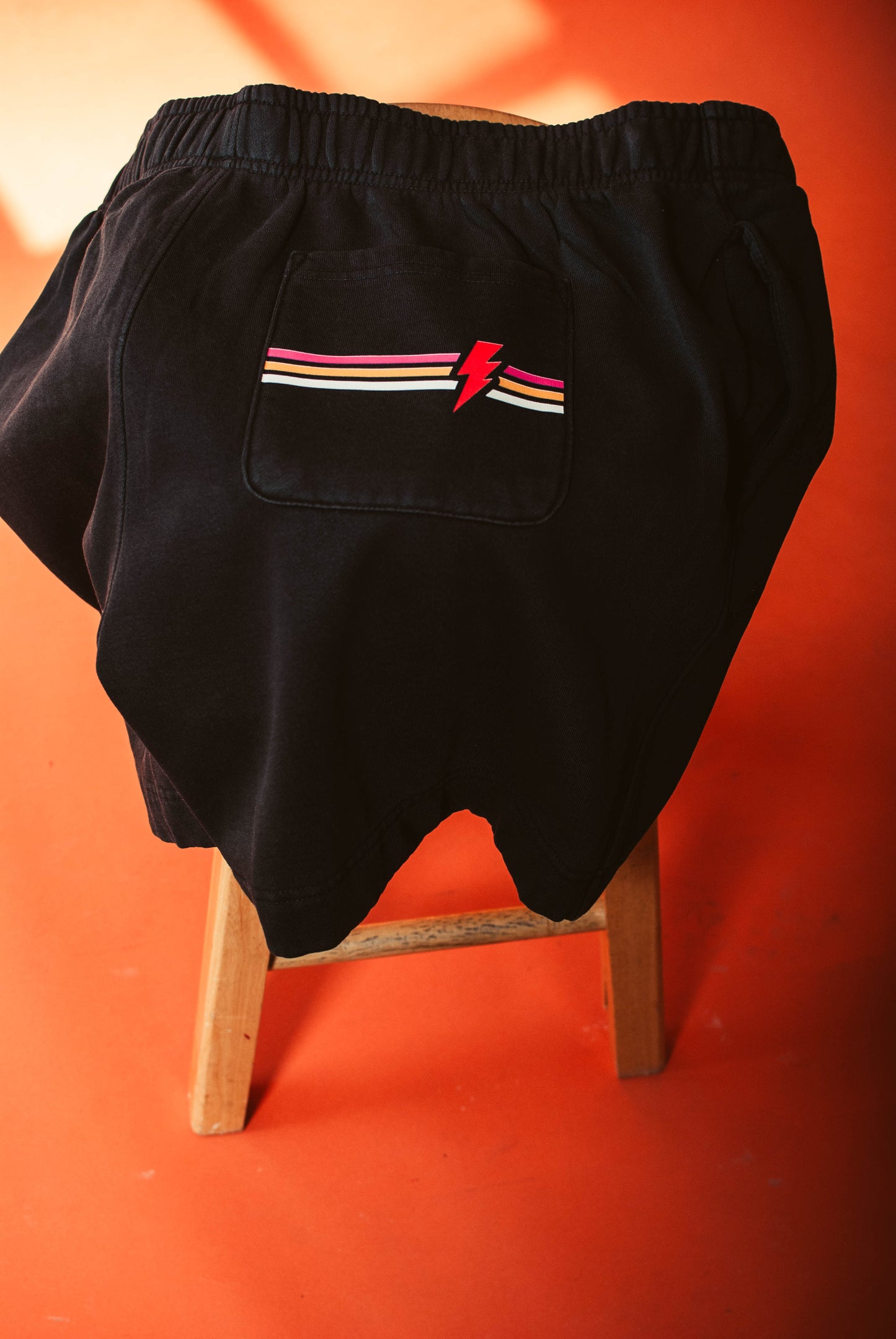 No apologies shorts with a colorful lightning bolt design on a wooden stool against an orange background