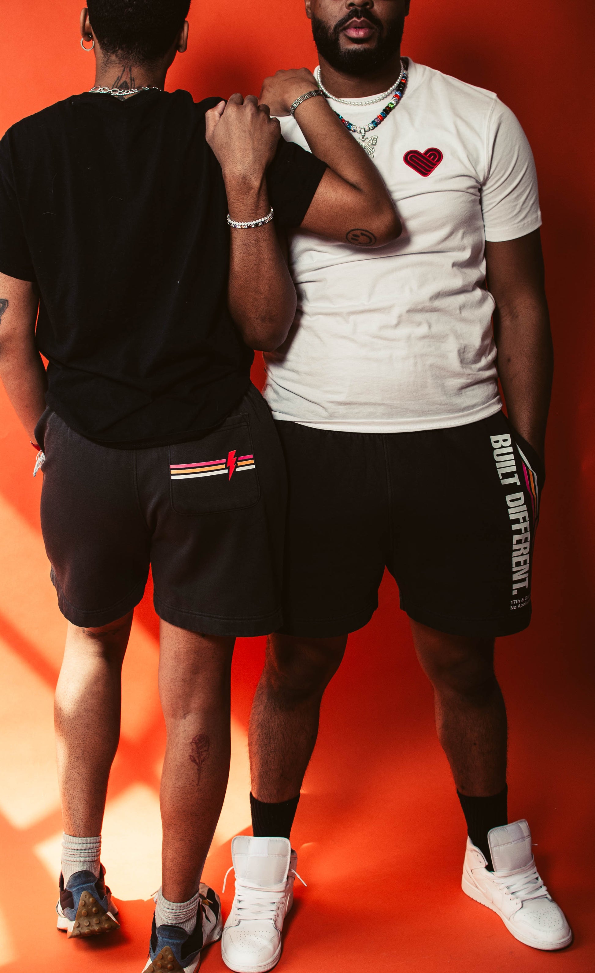 Two people standing against an orange background, wearing No Apologies shorts with visible branding.