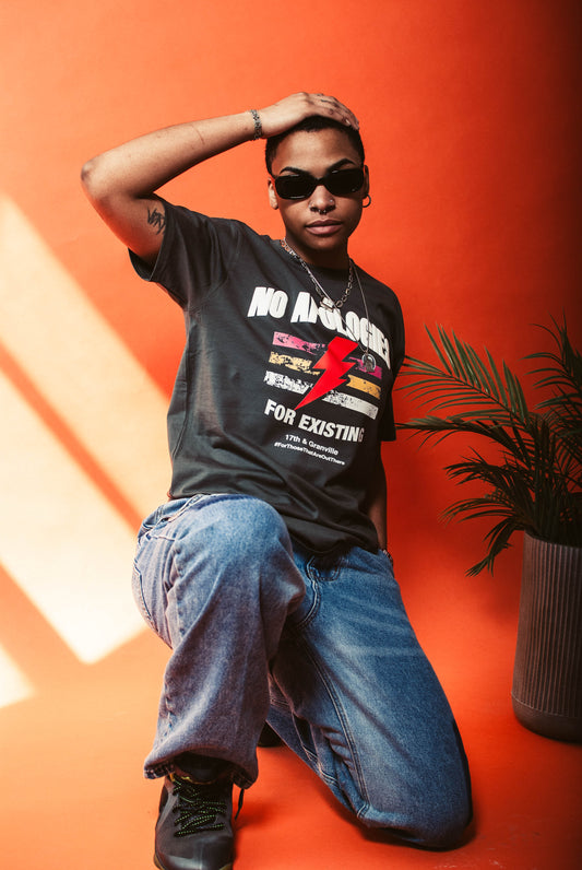 Person wearing a No Apologies t-shirt from 17th & Granville with graphic design and text, sitting on an orange background.