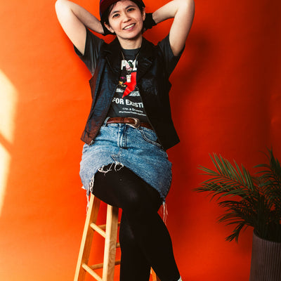 Person sitting on a stool against an orange wall wearing a No Apologies T-shirt from 17th & Granville 