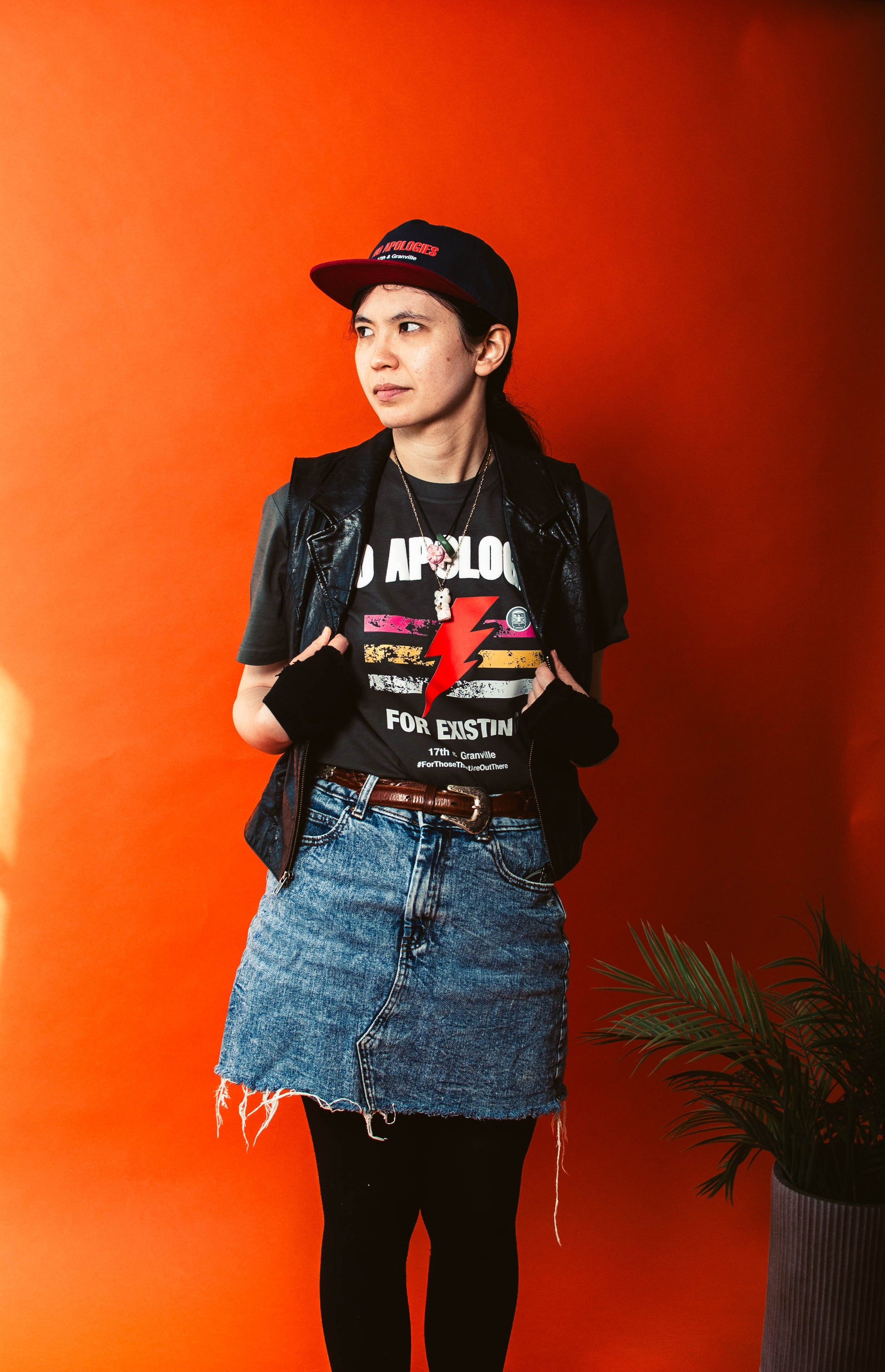 Person wearing a No Apologies t-shirt with colorful graphics and text, denim skirt, and black jacket against an orange wall.