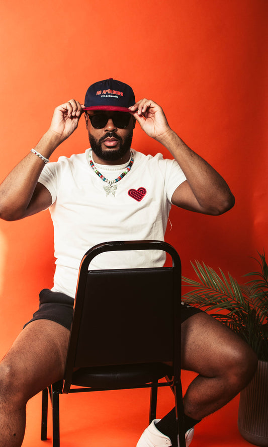 Man wearing a white t-shirt with a red heart, black shorts, and a No Apologies Hat from 17th & Granville sitting on a chair against an orange background.