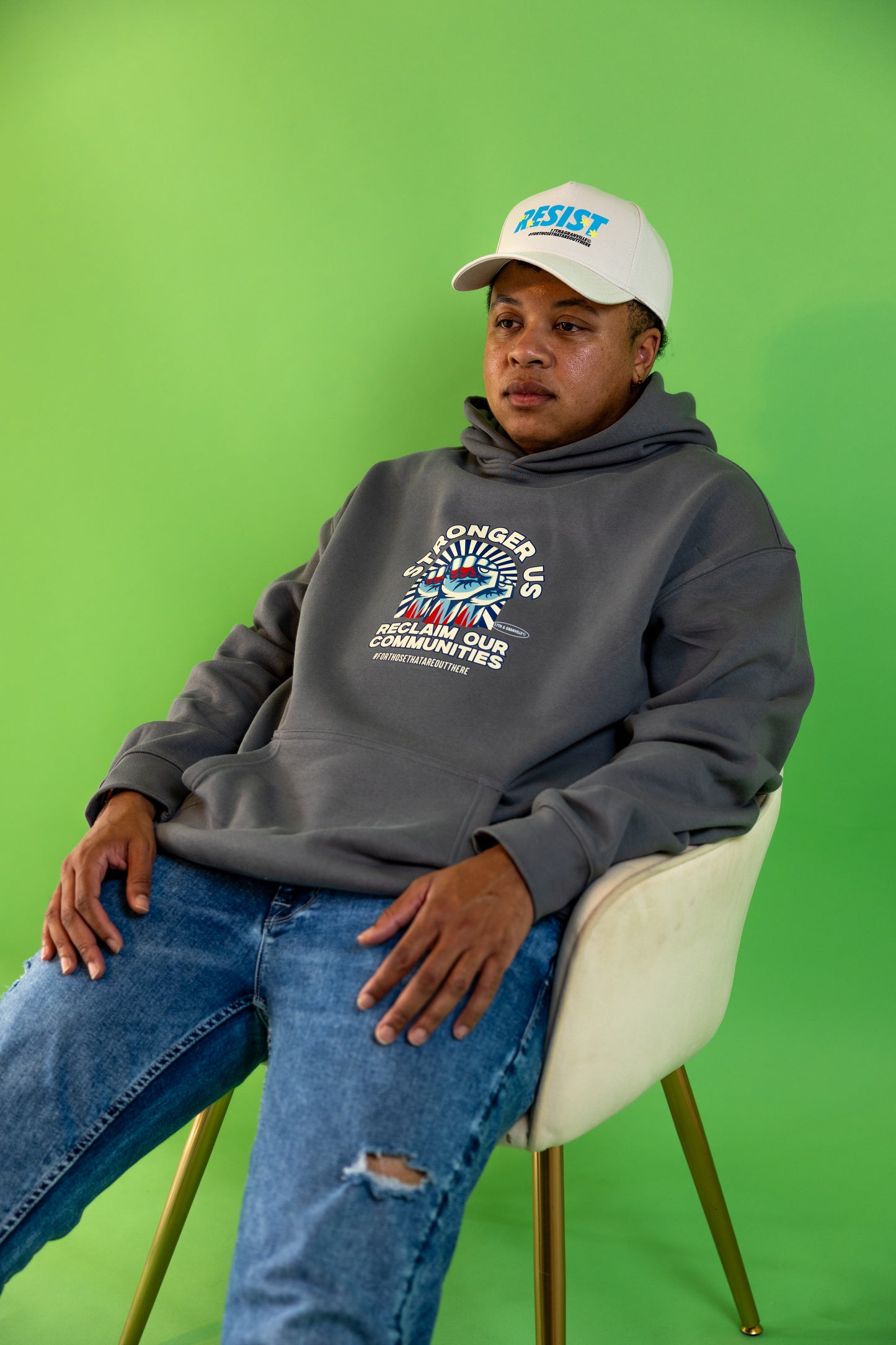 Person wearing a Reclaim Our Communities Hoodie by 17th & Granville – pullover fleece with bold raised-fist graphic and Stronger Us slogan, sitting on a chair against a green background