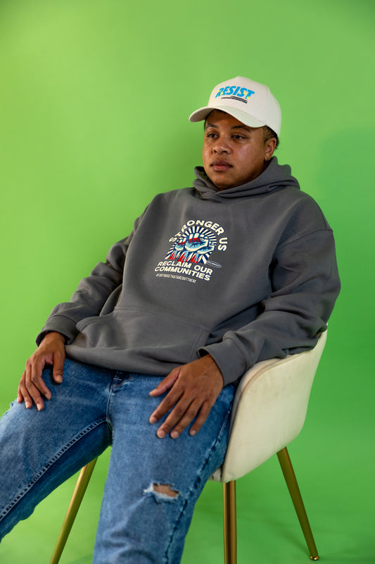 Person wearing a Reclaim Our Communities Hoodie by 17th & Granville – pullover fleece with bold raised-fist graphic and Stronger Us slogan, sitting on a chair against a green background