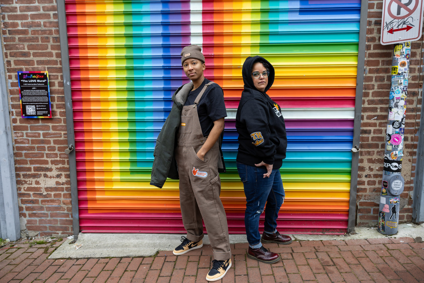 Two individuals stand back-to-back in front of a vibrant rainbow mural painted on a metal garage door. The person on the left wears brown overalls with a "Collective Action" patch, a black t-shirt, a brown beanie, and tan-and-black sneakers, with a green jacket draped over one shoulder. The person on the right wears a black hoodie with the "WE/US together" design on the front, blue jeans, and burgundy boots, with the hood up. They both have confident expressions, representing unity, empowerment, and communi