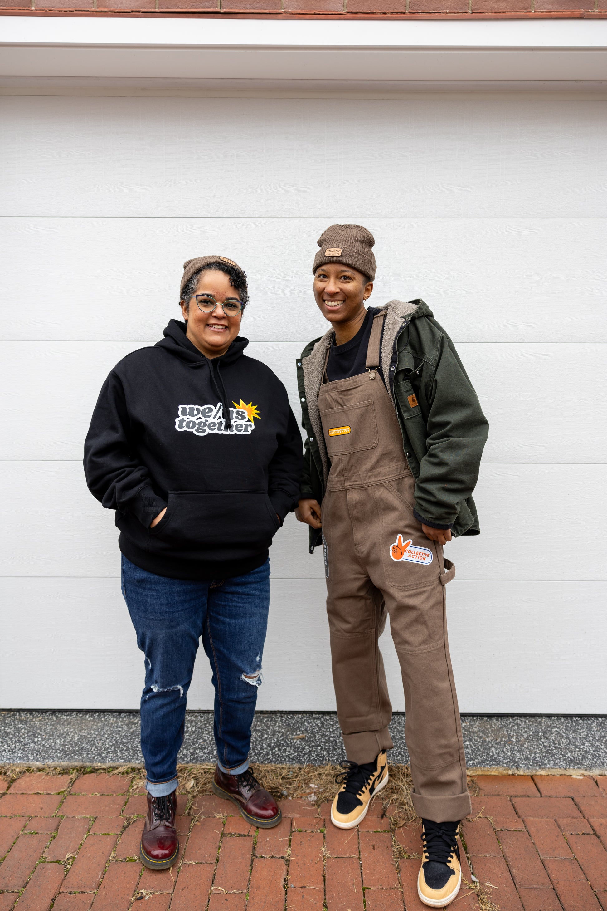 Two individuals stand side by side in front of a white garage door, smiling at the camera. The person on the left wears a black hoodie with the phrase "WE/US together" printed on the front, distressed blue jeans, a brown beanie, and round glasses. The person on the right wears brown overalls with a patch reading "Collective Action," a green jacket, a brown beanie, and tan-and-black sneakers. They both have their hands in their pockets and stand on a brick sidewalk.