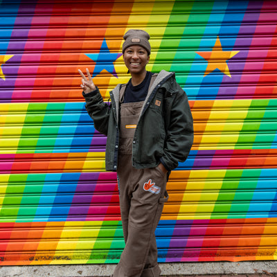 Model wearing brown We Us overalls with a We Us beanie in front of a rainbow background.